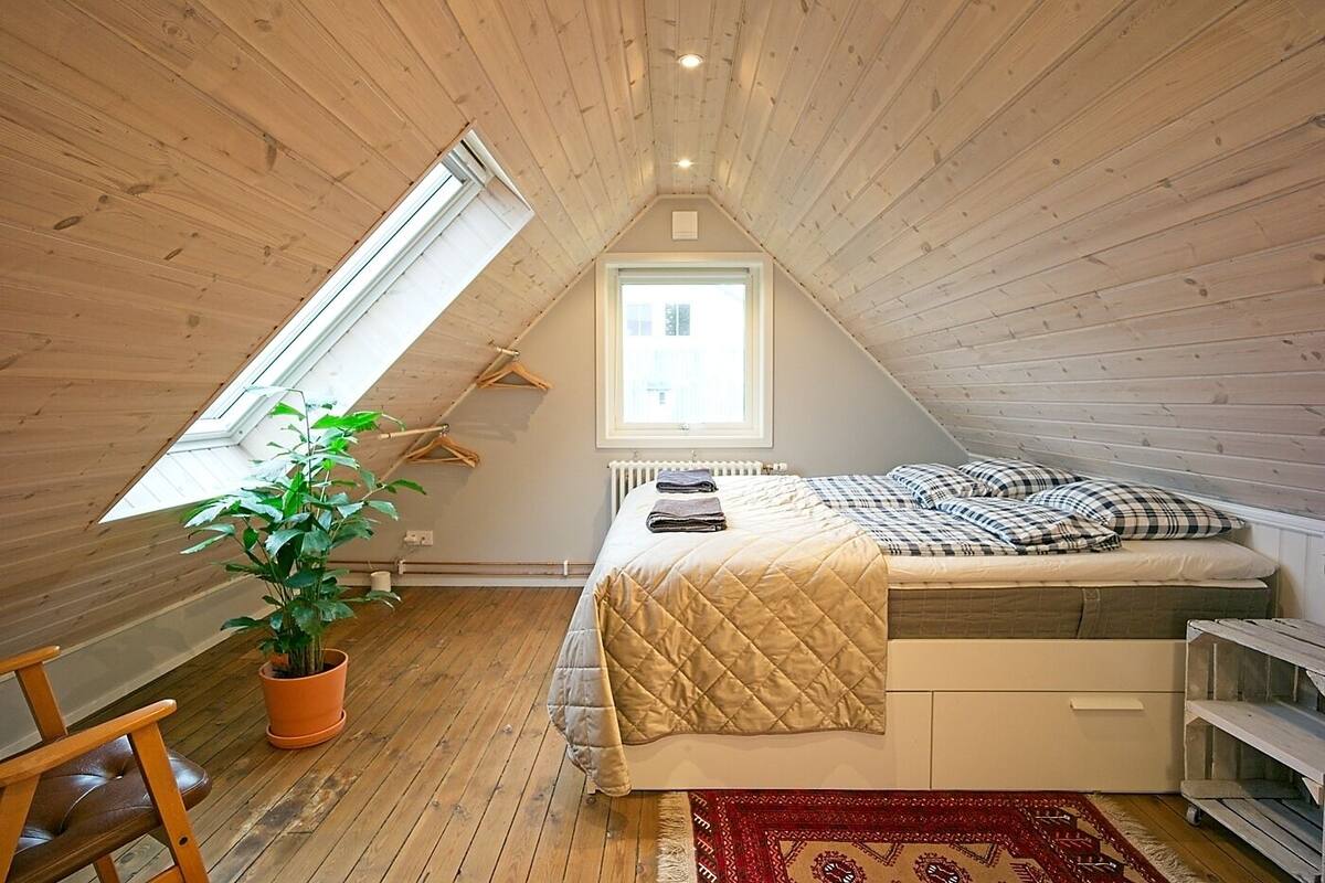 Ein kleines, gemütliches Schlafzimmer im Dachgeschoss mit schrägen Wänden, einem Oberlicht, einem Bett mit gesteppter Bettdecke, einer Topfpflanze und einem Heizkörper.