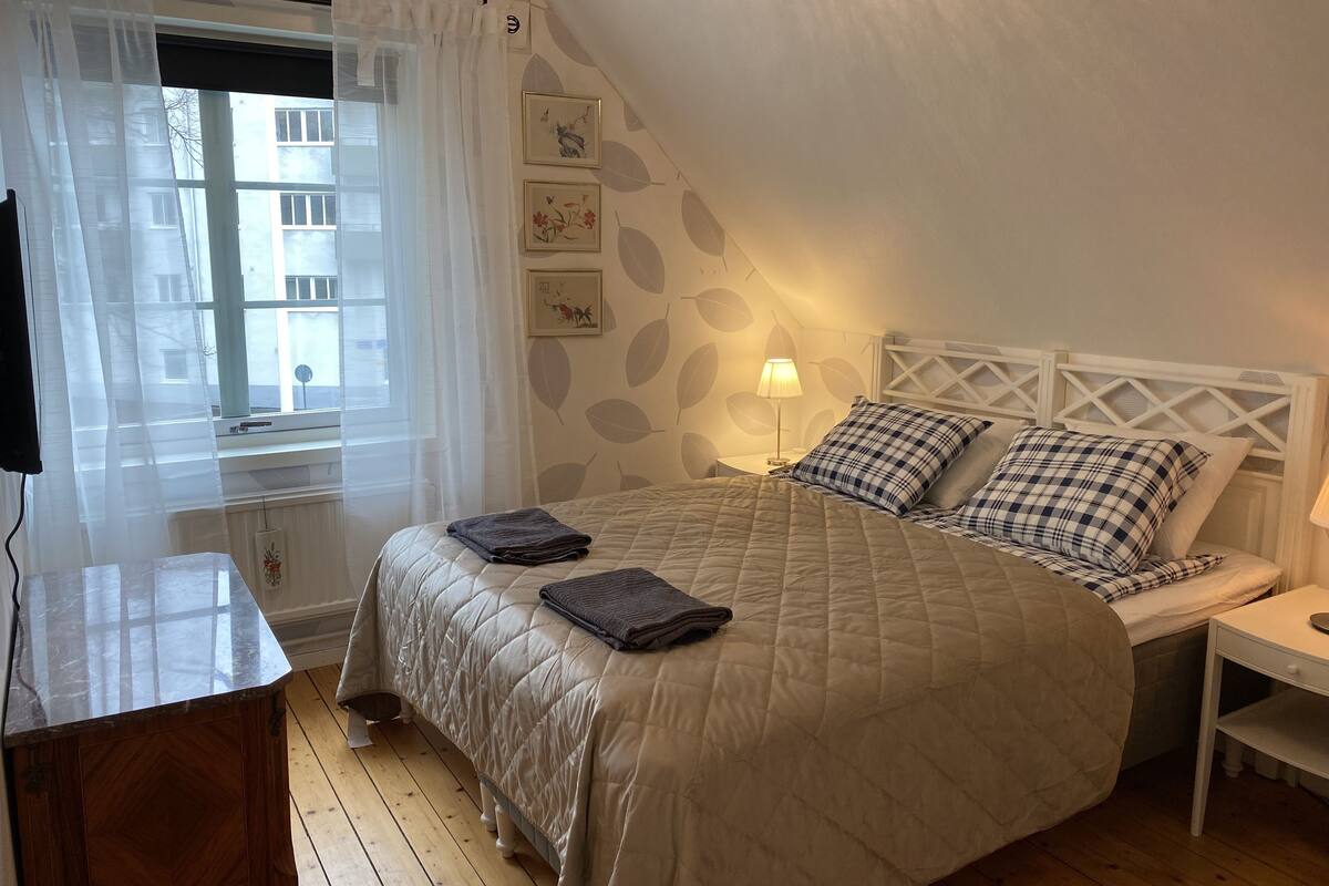 Ein Schlafzimmer mit einem Bett, einem Nachttisch, einer Lampe und einem Fenster mit Vorhängen.