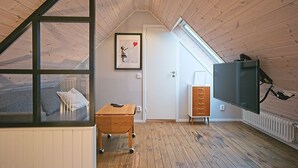 Une pièce avec un plafond en pente, un plancher en bois, une petite table en bois, une armoire blanche, un tableau encadré accroché au mur et un téléviseur fixé au mur.