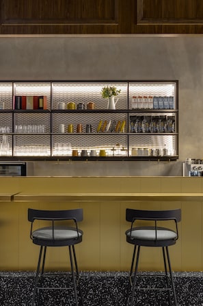 A bar area with a metal shelf displaying various items, two black chairs, and a yellow counter.