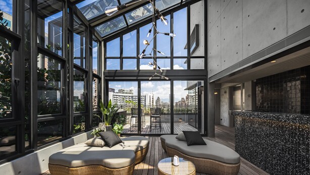 Een moderne rooftop lounge met rieten meubilair, grote ramen en uitzicht op de stad.