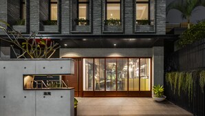 Een modern gebouw met een glazen ingang, een betonnen plantenbak met beplanting en een metalen hek met groen.