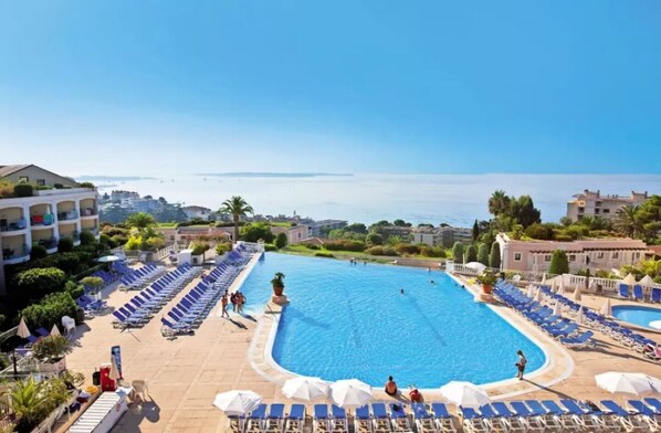 Une grande piscine à l’eau bleue, entourée de chaises longues et de parasols, avec des bâtiments et un ciel dégagé en arrière-plan.