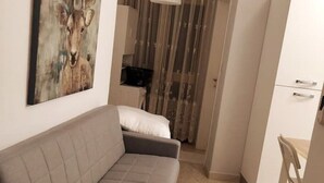 Ein modernes Wohnzimmer mit einem grauen Sofa, einem Bild eines Rehs, einer weißen Pendelleuchte und Blick in ein Schlafzimmer mit Fenster und Vorhängen.