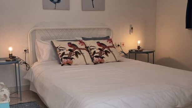 Ein ordentlich bezogenes Bett mit floralen Kissen, ein Nachttisch mit Lampe und zwei an der Wand hängende Bilder.