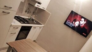 Eine kleine Küche mit Holztisch und -stühlen, einem eingebauten Backofen und einem an der Wand befestigten Fernseher, der die Serie 'Lucifer' zeigt.
