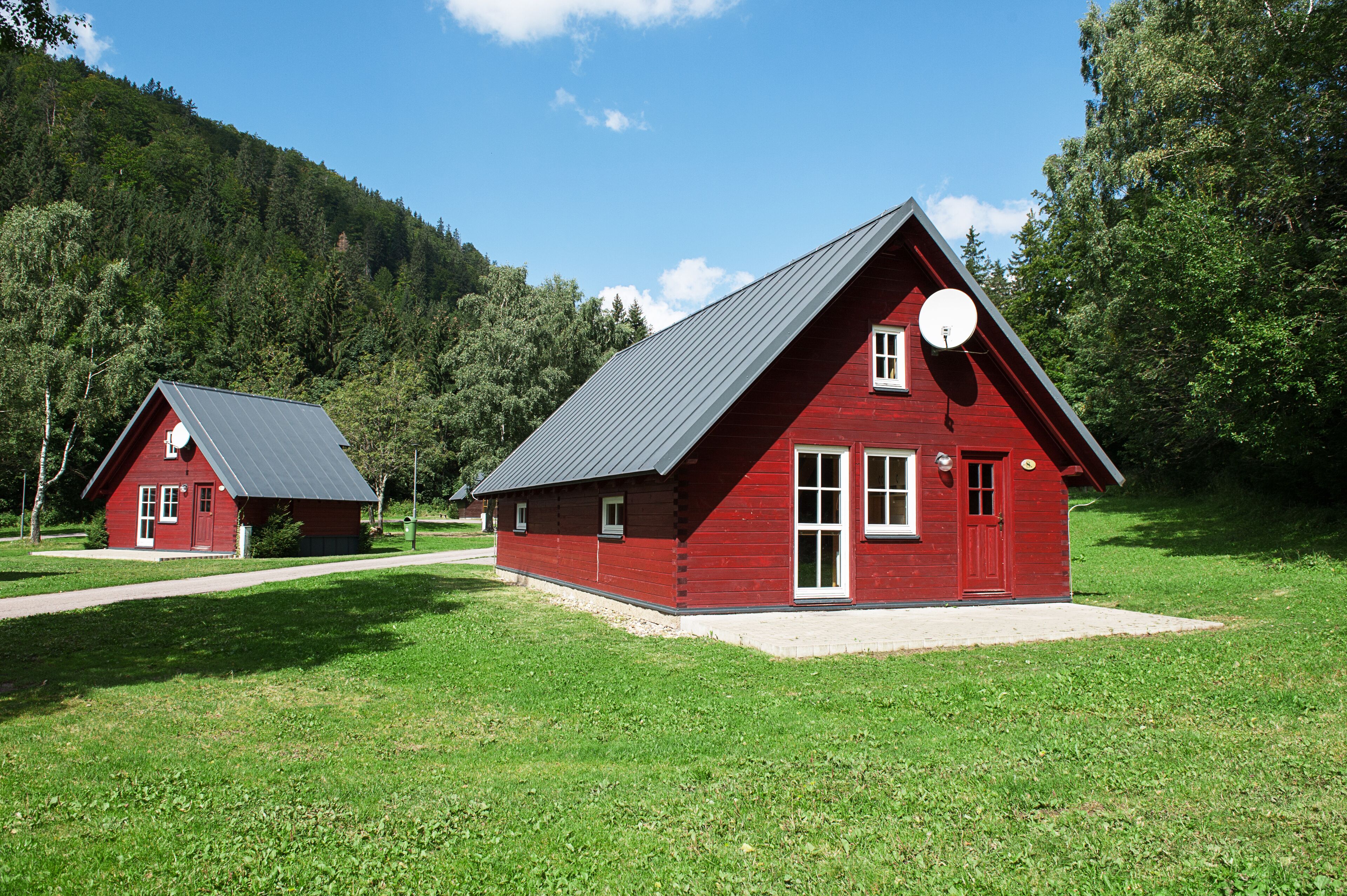 Due cabine di legno rosse con tetti spioventi, immerse in un prato con una collina boscosa sullo sfondo.