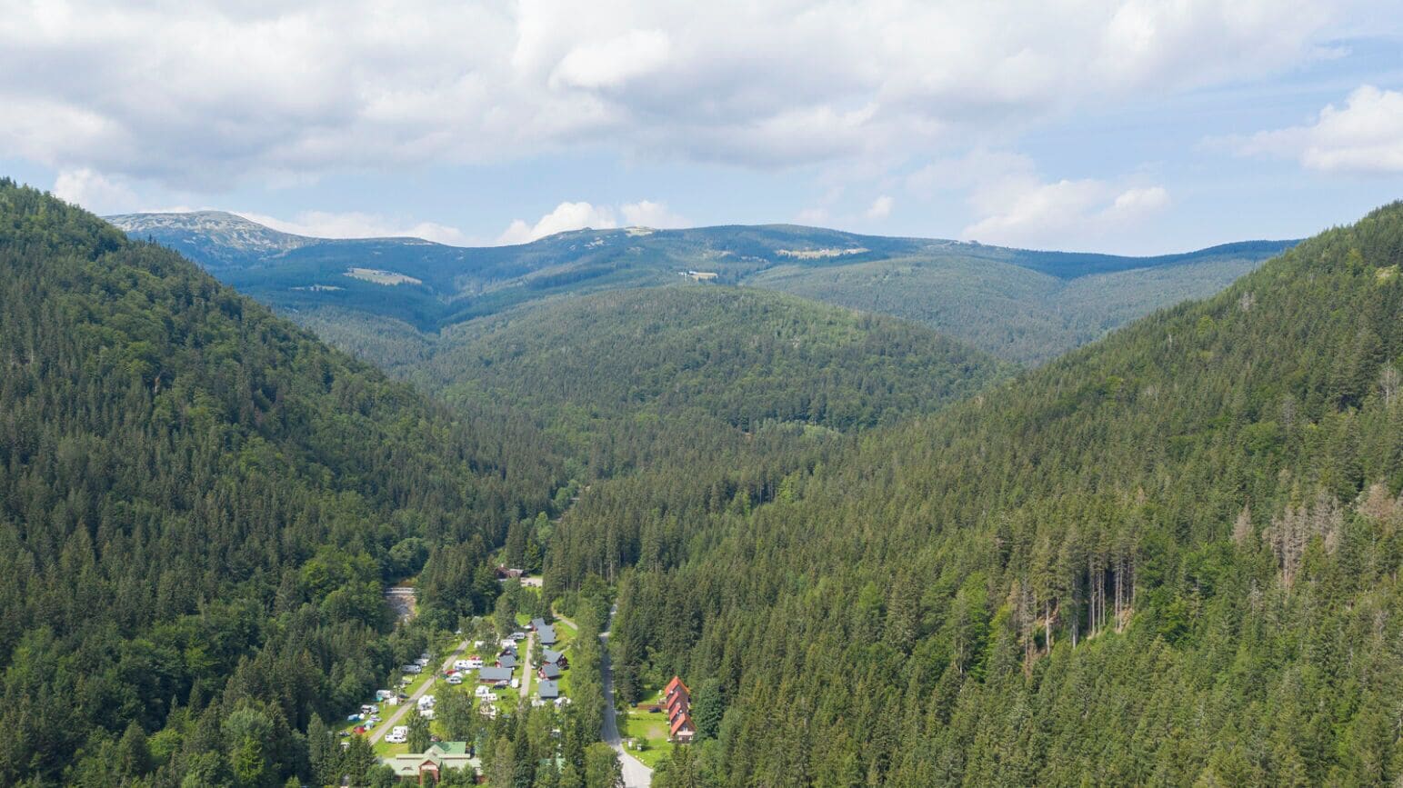 A forested valley with a small cluster of buildings, likely a resort or lodge, surrounded by dense trees and distant mountains.