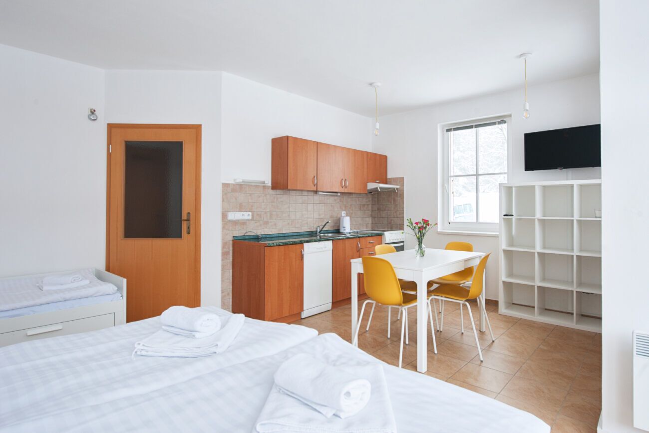 A modern hotel room with a bed, a small kitchenette, a dining area with a table and chairs, and a TV mounted on the wall.