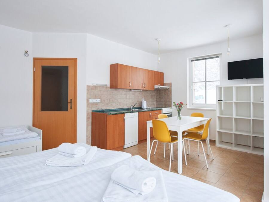 A modern hotel room with a bed, a small kitchenette, a dining area with a table and chairs, and a TV mounted on the wall.