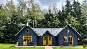A two-story cabin with a dark blue exterior and yellow trim, surrounded by lush greenery and trees. - Base Camp Medvědín (Spindleruv Mlyn)