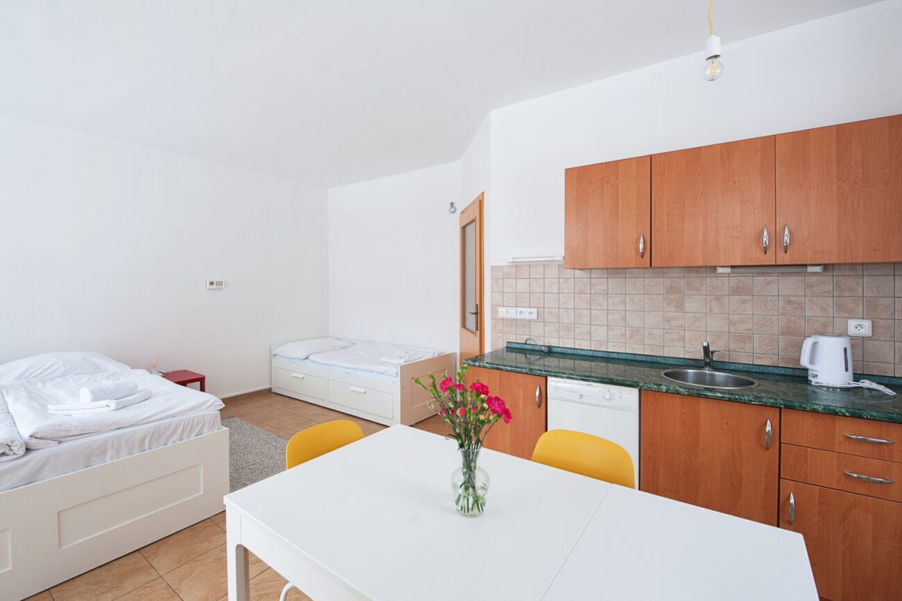 Een kleine hotelkamer met een bed, een tafeltje met een bloevas, een kitchenette met houten kastjes en een witte wasbak.