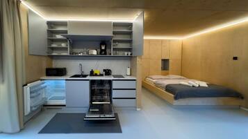 Un dormitorio compacto con kitchenette integrado, una cama y un pequeño espacio de trabajo.