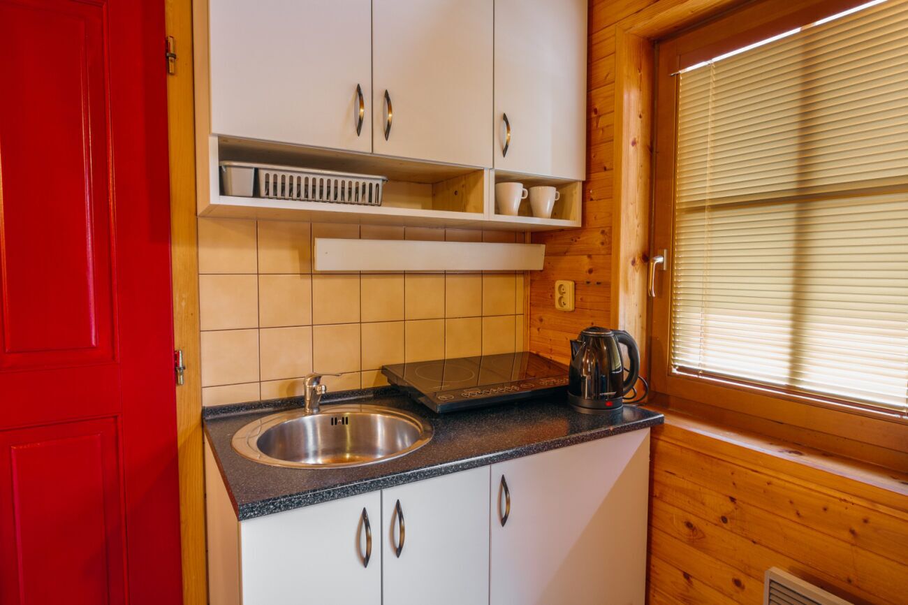 Een kleine keuken met witte kastjes, een RVS spoelbak, een zwarte waterkoker en een raam met zonwering.