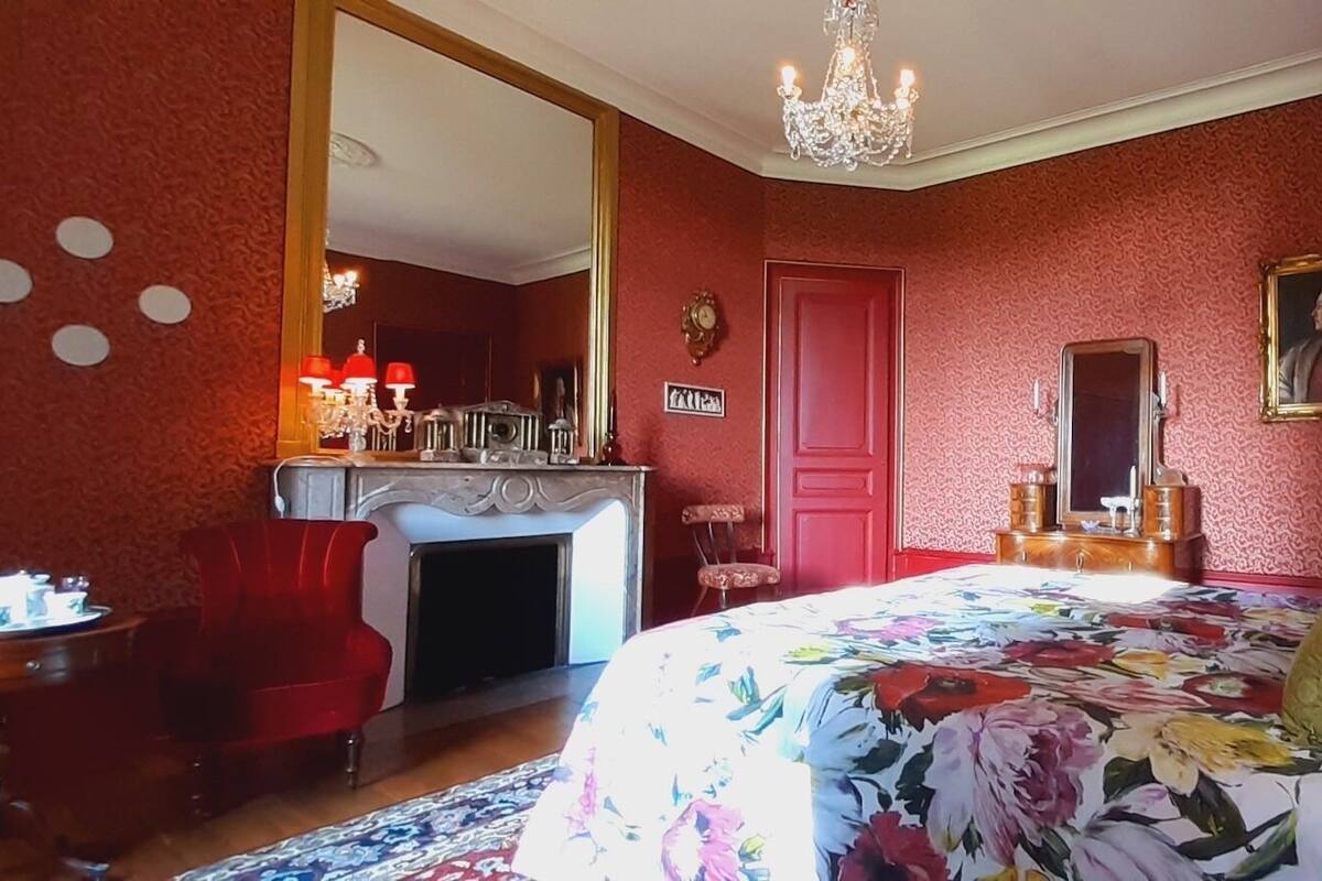 A bedroom with a floral bedspread, a red chair, a dresser, a mirror, and a chandelier.