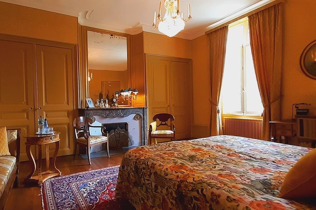 A hotel room with a large bed, a sitting area with a chair and a small table, a fireplace, and a large mirror on the wall.