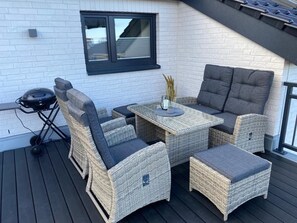 Un espace extérieur aménagé avec des meubles en osier, comprenant une table et des chaises, un barbecue et une fenêtre équipée de stores.