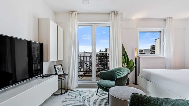 Une chambre d’hôtel moderne dotée d’un grand téléviseur à écran plat, d’un fauteuil vert confortable, d’un canapé et d’une grande fenêtre donnant sur la ville.