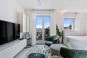 Habitación de hotel moderna con un televisor de pantalla plana grande, un sillón verde confortable, un sofá y un ventanal con vistas a la ciudad.
