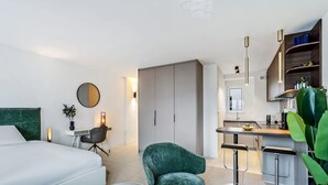 Une chambre d’hôtel moderne équipée d’un lit, d’un bureau, d’un fauteuil vert, d’un petit coin cuisine et d’une grande plante.