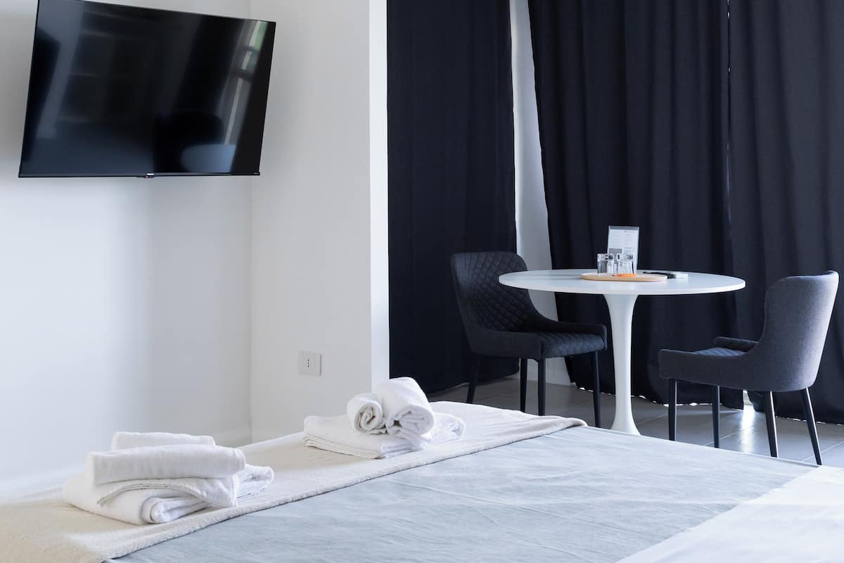 A modern hotel room with a bed, a TV mounted on the wall, a small round table with chairs, and a black curtain.