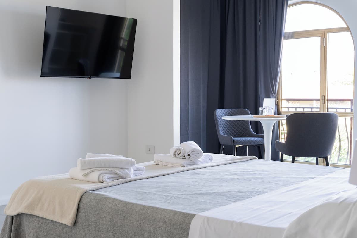 A hotel room with a bed, a flat-screen TV mounted on the wall, a desk with a chair, and a large window with curtains.