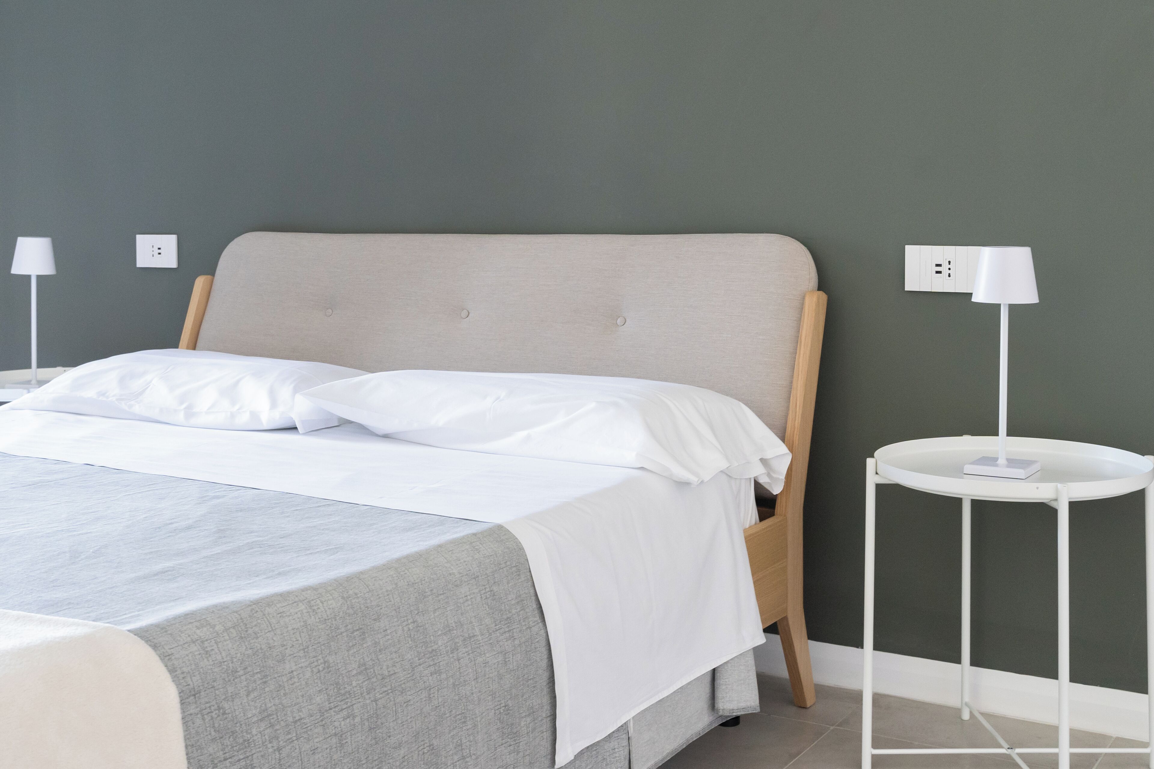 Ein ordentlich bezogenes Bett mit weißen Leinen, einer grauen Tagesdecke und einem hölzernen Kopfteil. Ein weißer Nachttisch mit Lampe und Steckdose ist vorhanden. Die Wand ist grün gestrichen.