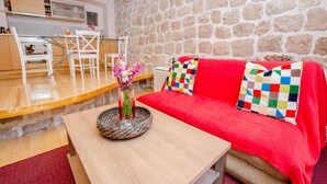 Une pièce chaleureuse avec un mur en pierre, une table en bois ornée d’un vase de fleurs, un canapé rouge avec un coussin coloré, et un coin cuisine à l’arrière-plan.