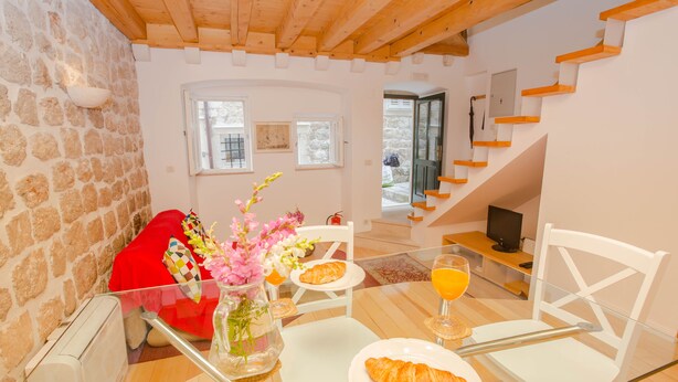 Un coin repas avec une table mise pour le petit-déjeuner, comprenant des croissants et un verre de jus d’orange. On y trouve des chaises blanches et une chaise rouge, avec un mur en pierre et des poutres en bois en arrière-plan.