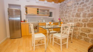 Une cuisine avec des meubles en bois, un réfrigérateur en acier inoxydable, une table à manger en verre avec des chaises blanches et un mur en pierre.
