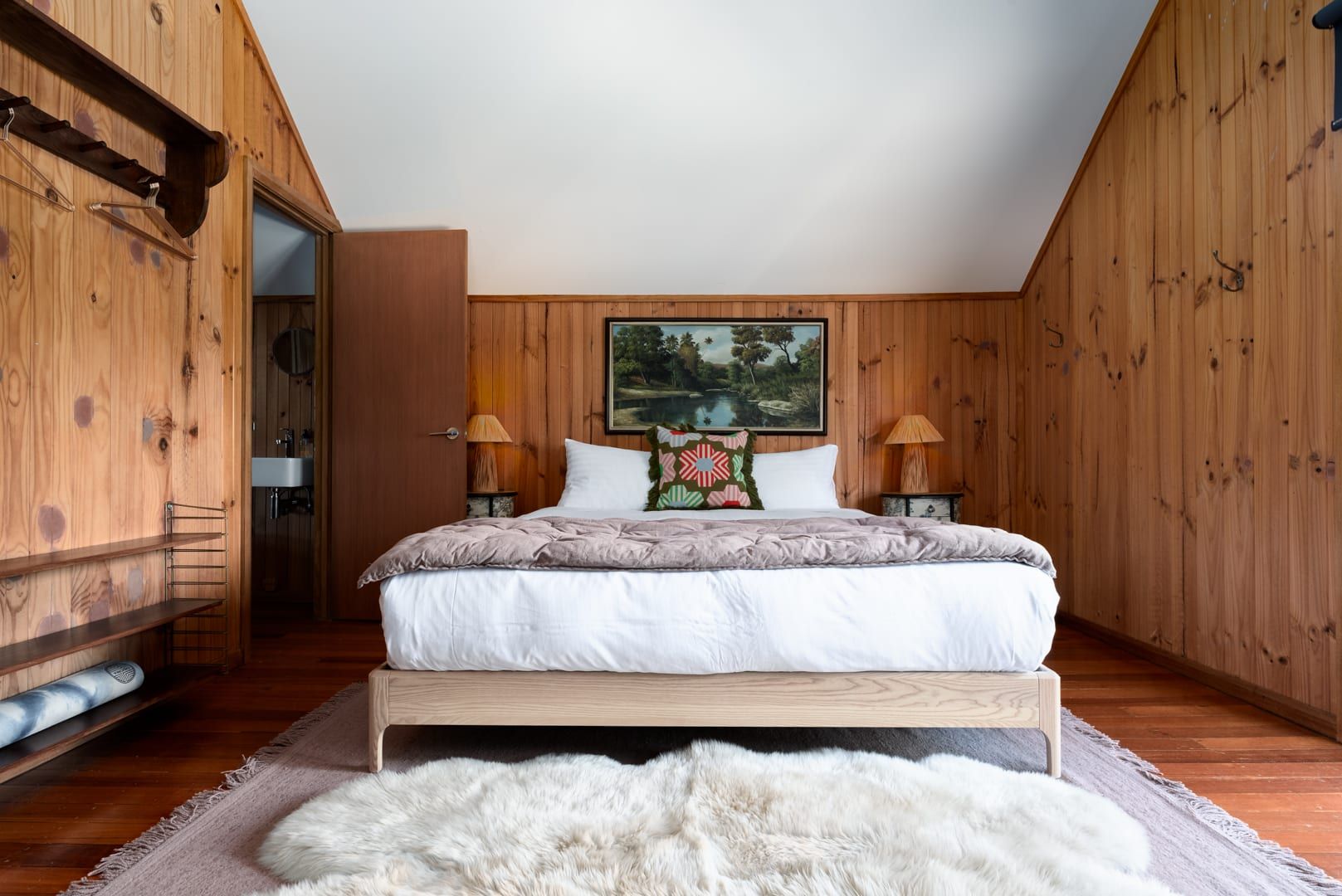 Ein Schlafzimmer mit Holzbalkendecke, einem Bett mit weißer Bettwäsche, zwei Nachttischlampen, einem Bild an der Wand und einem Fellteppich auf dem Boden.