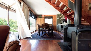 Interior acolhedor de uma cabana, com área de jantar, piso de madeira, lareira e vista através de grandes janelas.