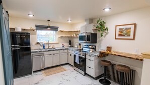 A modern kitchen with a black refrigerator, stainless steel appliances, a microwave, and a built-in oven. There are two bar stools and a potted plant. - Dollhouse Downtown (Fairbanks)