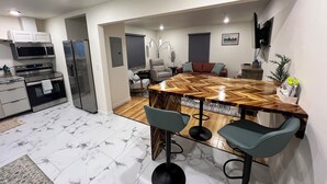 A modern kitchen with built-in appliances, a dining area with a wooden table and bar stools, and a living space with a sofa and a TV. - Dollhouse Downtown (Fairbanks)