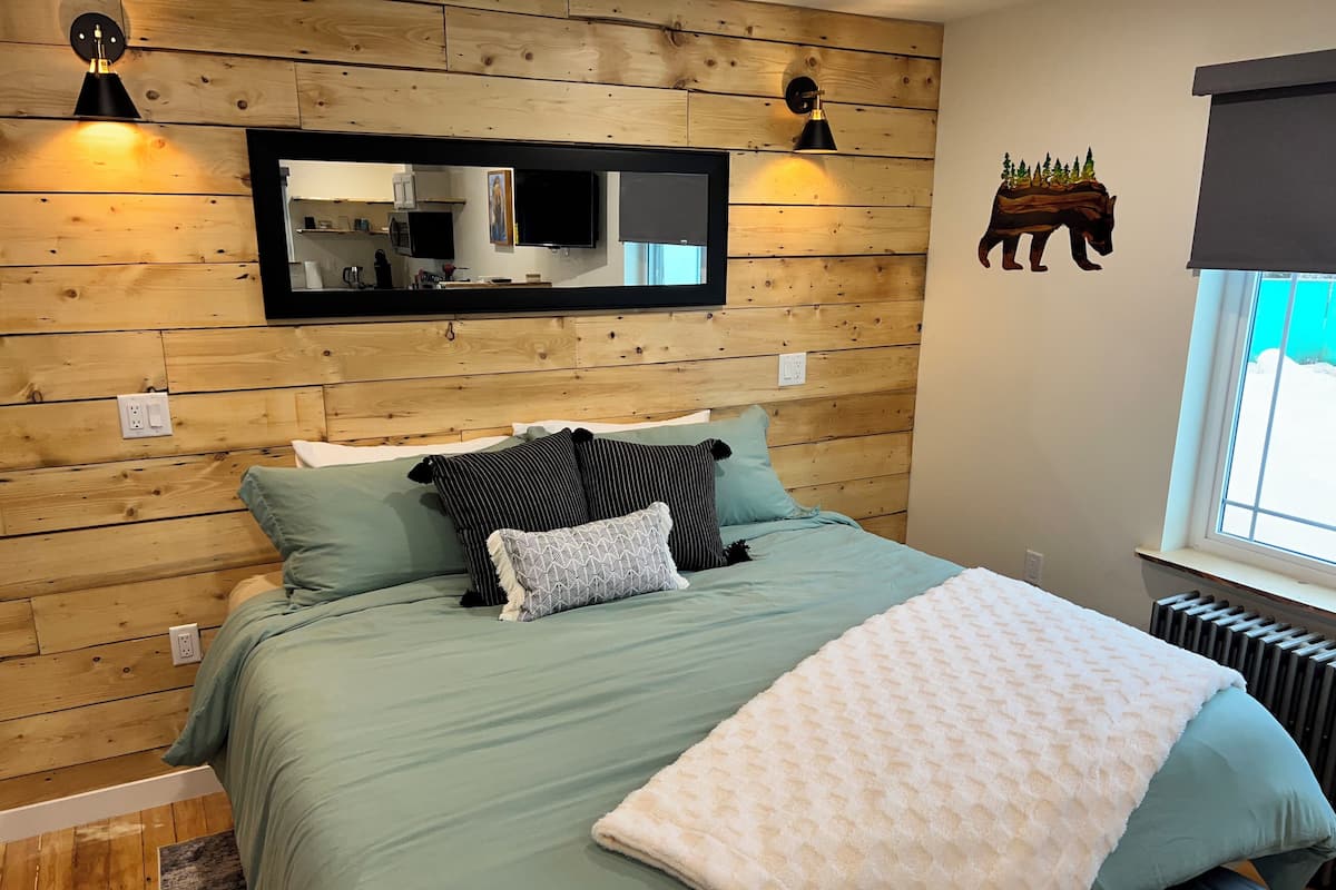 A bedroom with a wooden wall panel, a bed with green bedding, two bedside lamps, a large mirror, and a window with a view of trees.