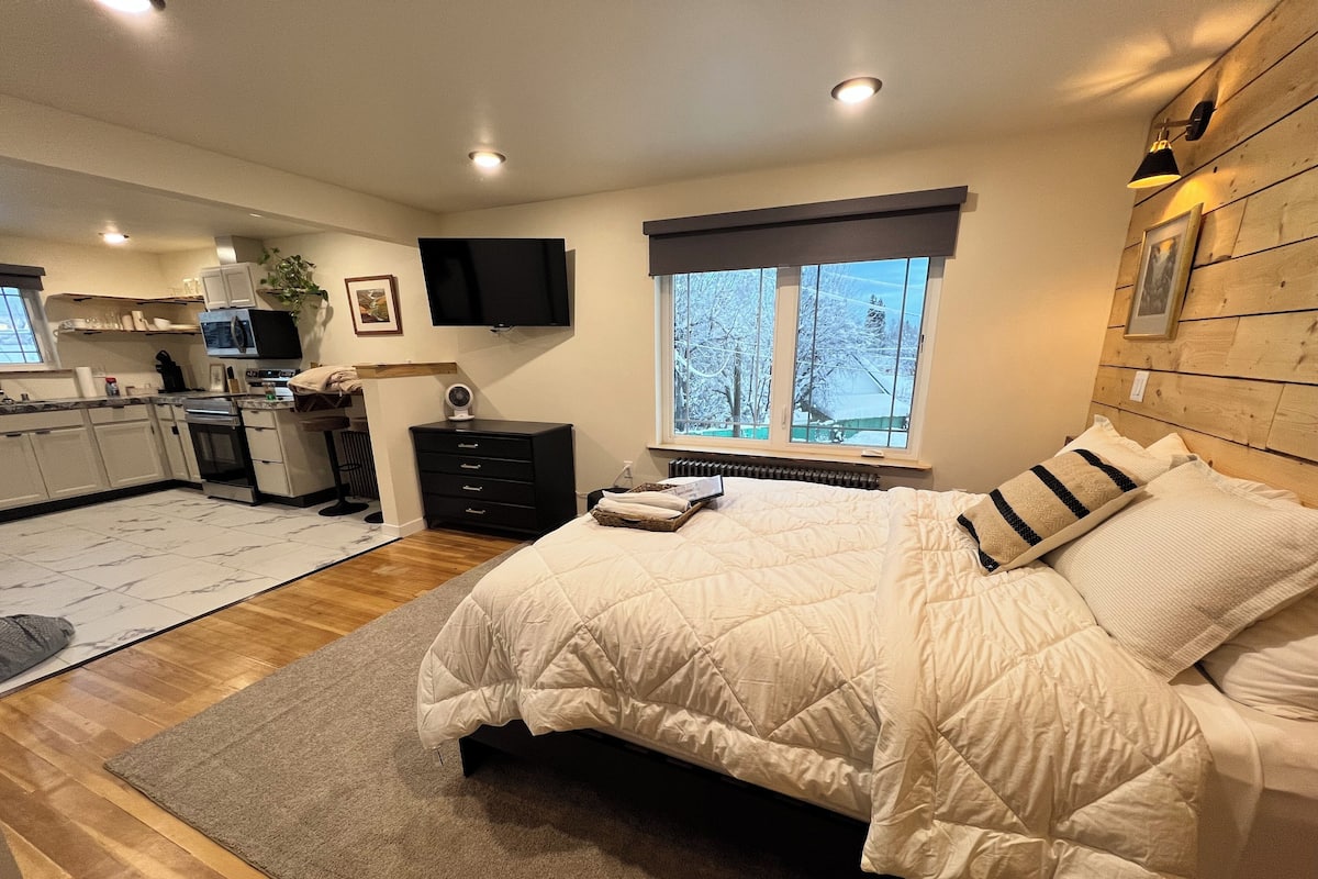 A bedroom with a large bed, a TV, a window with a view of trees, and a kitchen in the background.