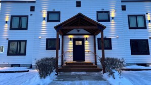 A white building with a dark entrance door, multiple windows, and a small porch with lighting. - Dollhouse Downtown (Fairbanks)