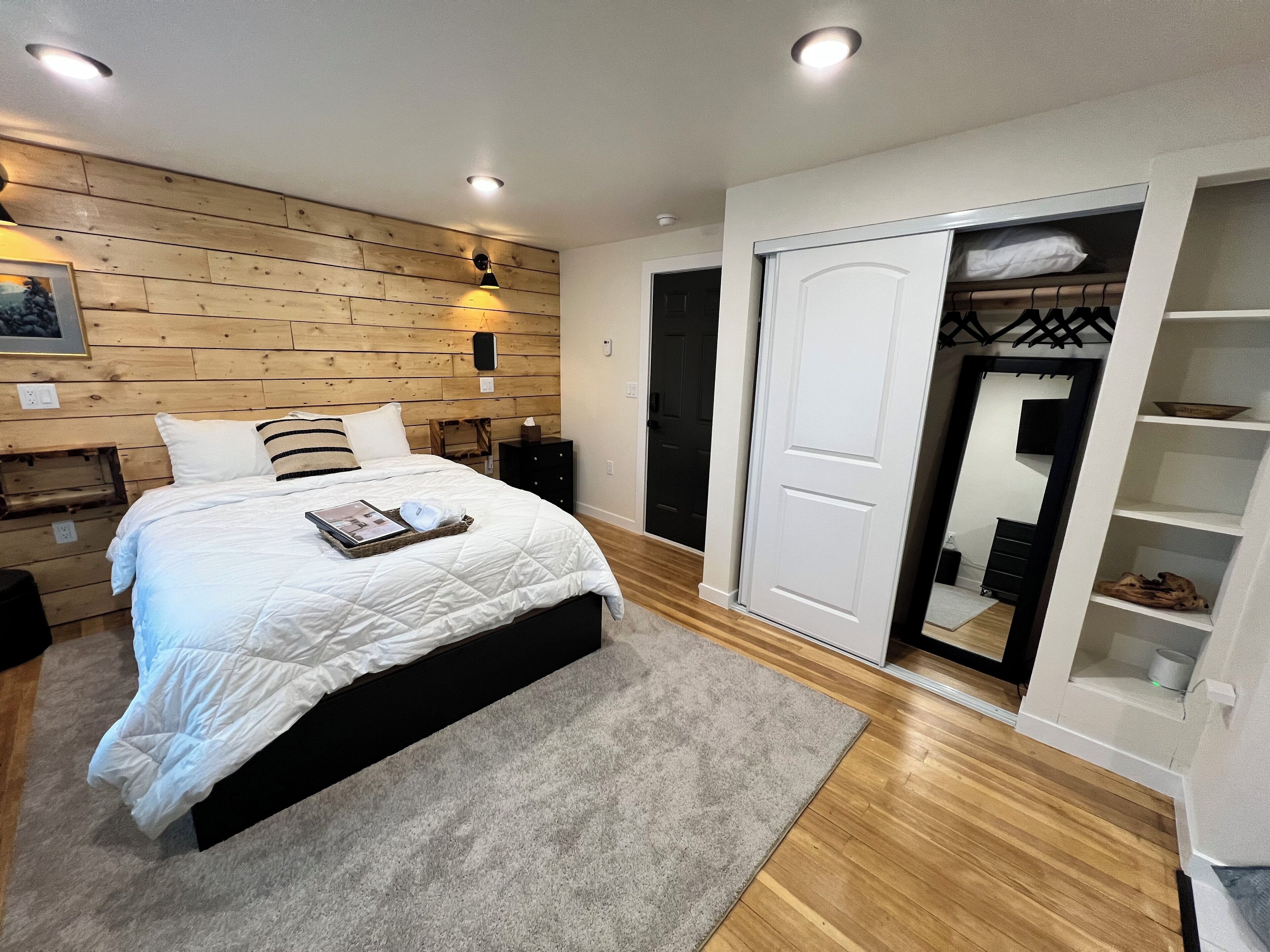 Une chambre avec un mur en bois, un lit avec une literie blanche, un tapis gris, une armoire blanche et un miroir.