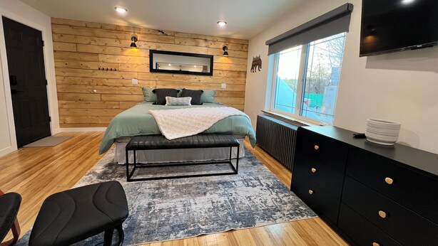 A bedroom with a bed, bench, desk, and TV. - Dollhouse Downtown (Fairbanks)