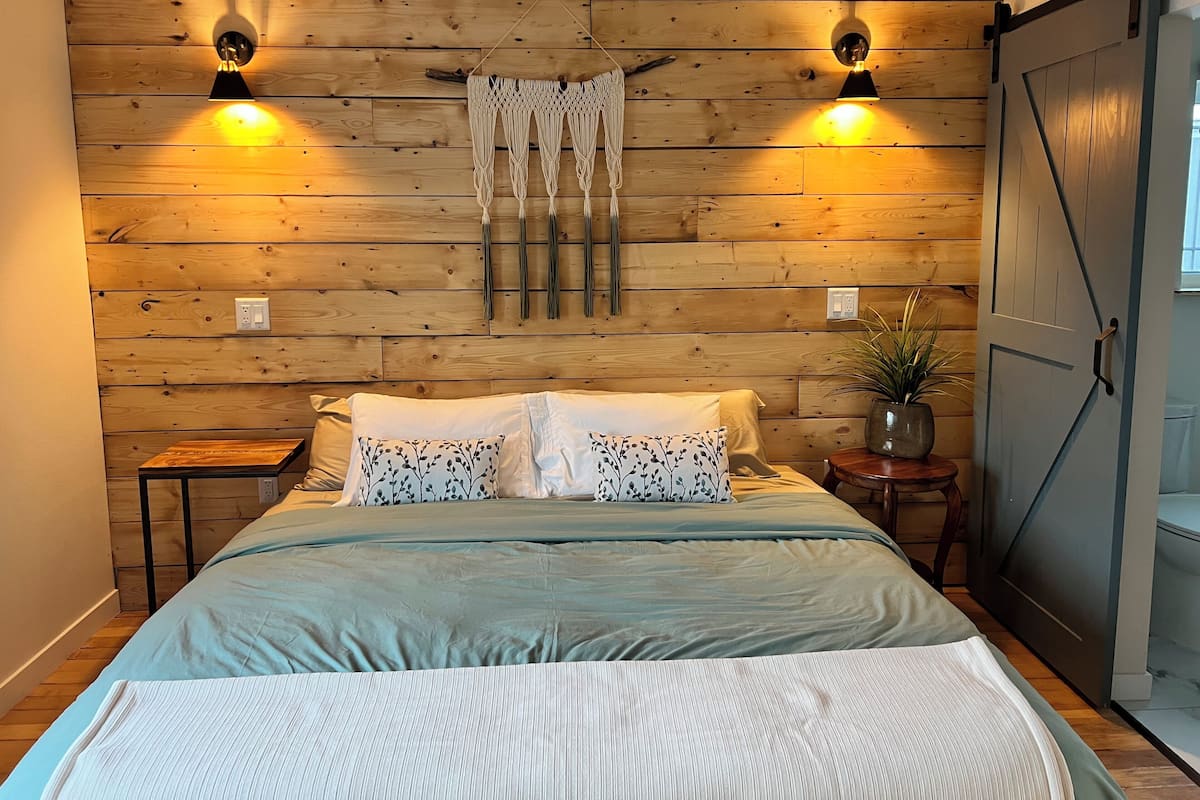 A bedroom with a wooden wall, two wall-mounted lights, a macrame wall hanging, a bed with two pillows, a small table, and a potted plant.