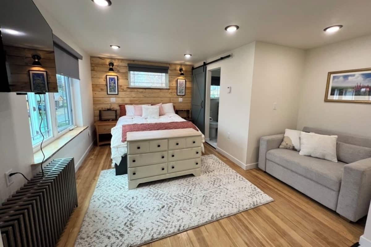 A modern bedroom with a large bed, a dresser, a gray sofa, and a window with wooden paneling.