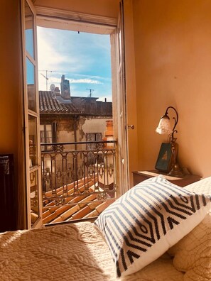 A bedroom with a bed, a patterned pillow, a nightstand with a lamp, and a view of a balcony with a railing and buildings in the background. - Amazing full furnished apartment in a very central location. (Arles)