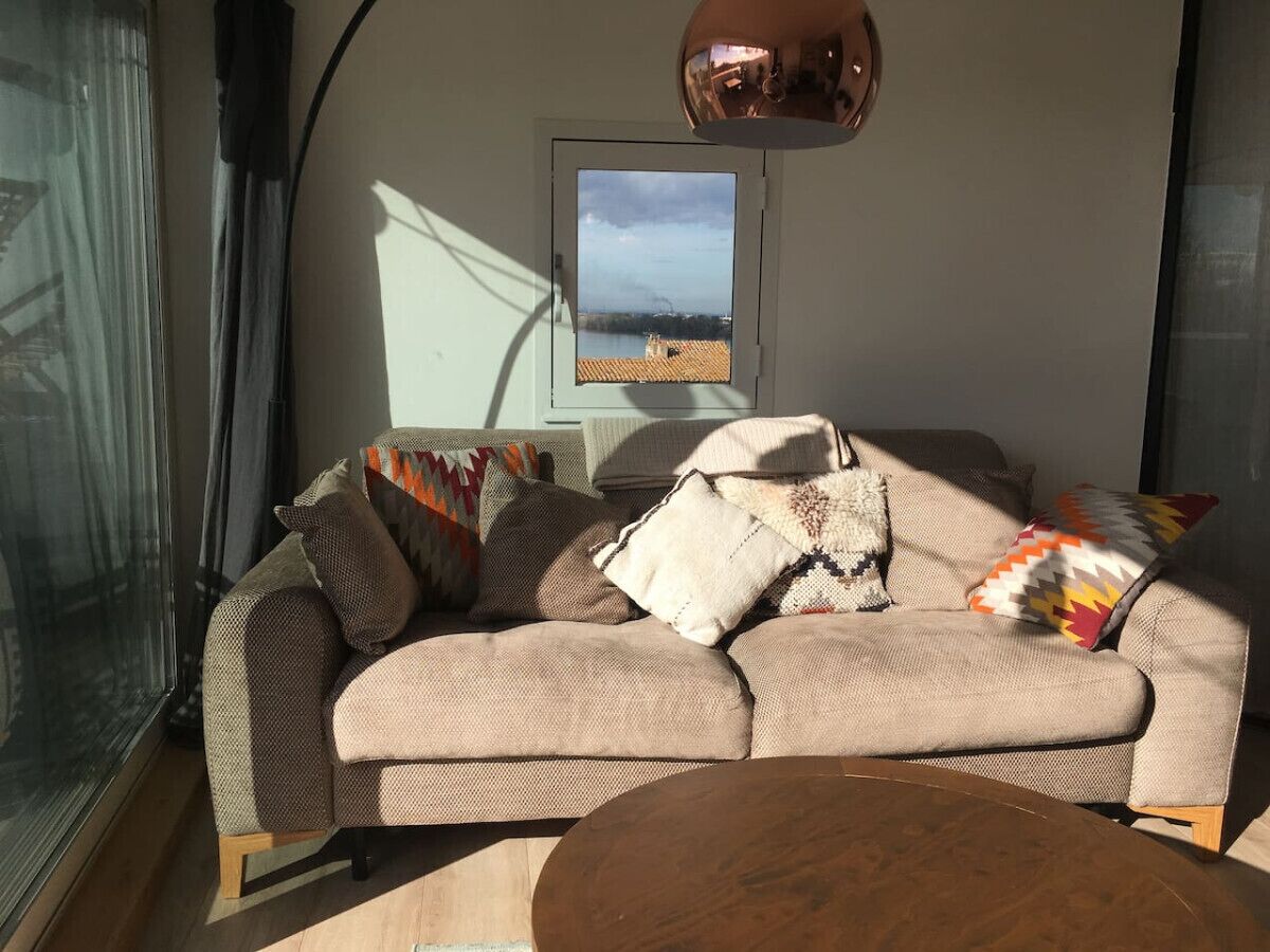 A modern living room with a beige sectional sofa, multiple patterned throw pillows, a wooden coffee table, and a view of a body of water through a window.