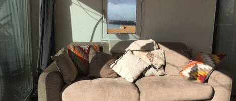 A modern living room with a beige sectional sofa, multiple patterned throw pillows, a wooden coffee table, and a view of a body of water through a window.