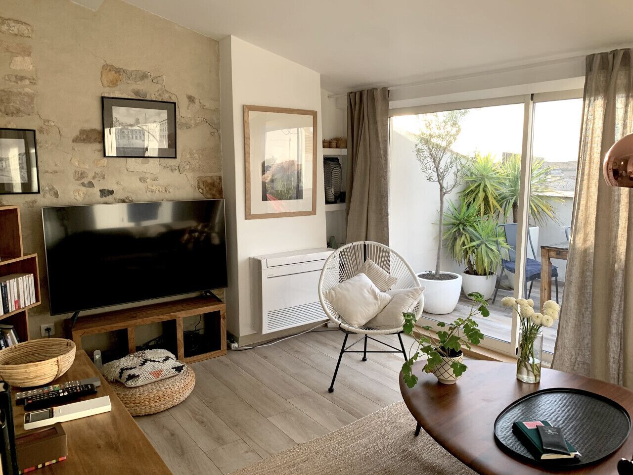 Sala de estar com televisão, uma cadeira branca, mesa de madeira e varanda com plantas.