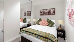 Un dormitorio moderno con una cama grande, un espejo, dos mesitas de noche con lámparas y una obra de arte floral en la pared.