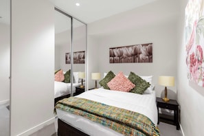 A modern bedroom with a large bed, a mirror, two bedside tables with lamps, and a floral artwork on the wall.