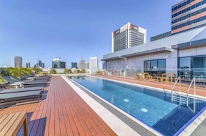 A rooftop swimming pool with sun loungers and a city skyline in the background.