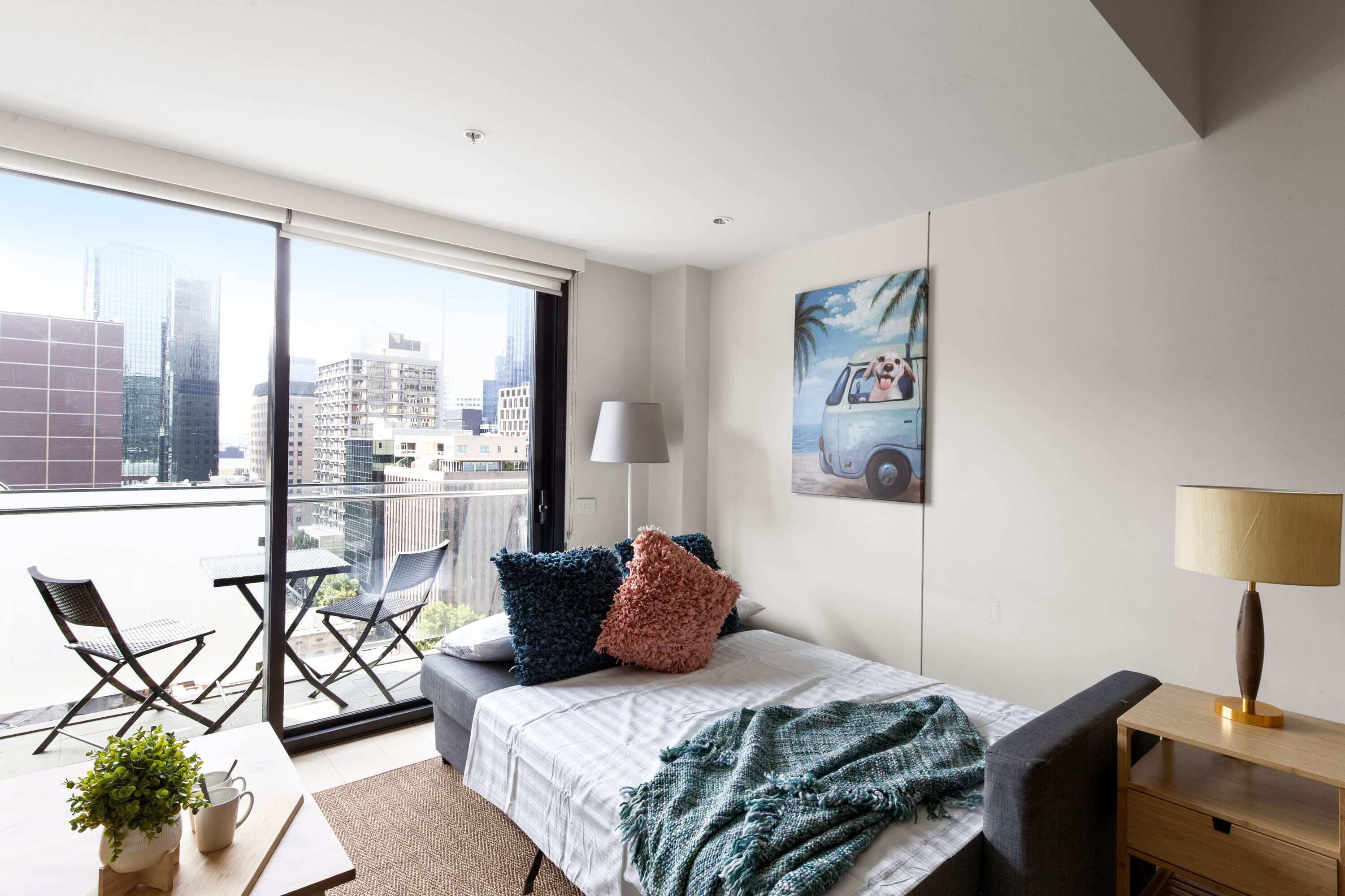 A modern hotel room with a large balcony, a bed with a green and white blanket, and a city view.