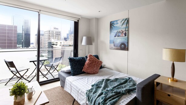Una habitación de hotel moderna con un balcón grande, una cama con manta verde y blanca, y vistas a la ciudad.